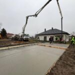 Na zdjęciu widać plac budowy z betonowaną powierzchnią. Ludzie w odblaskowych ubraniach, ciężarówka i dom w tle. Dzień jest pochmurny.