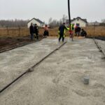 Grupa pracowników buduje fundamenty z betonu. Noszą odzież ochronną, a w tle są domy i pola. Niebo jest szare i zachmurzone.