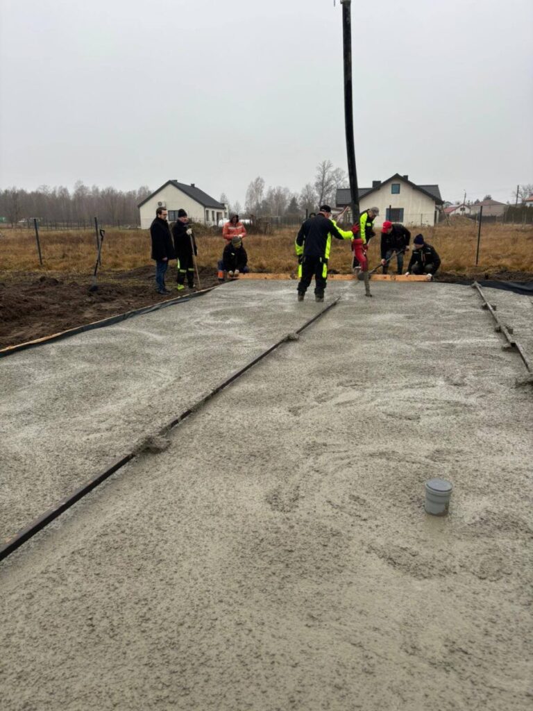 Grupa pracowników buduje fundamenty z betonu. Noszą odzież ochronną, a w tle są domy i pola. Niebo jest szare i zachmurzone.