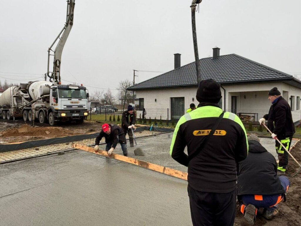Czterech pracowników w kurtkach wylewa beton przed domem z czarnym dachem. W tle widać ciężarówkę z betoniarką. Dzień jest pochmurny.