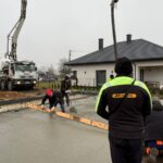 Czterech pracowników w kurtkach wylewa beton przed domem z czarnym dachem. W tle widać ciężarówkę z betoniarką. Dzień jest pochmurny.