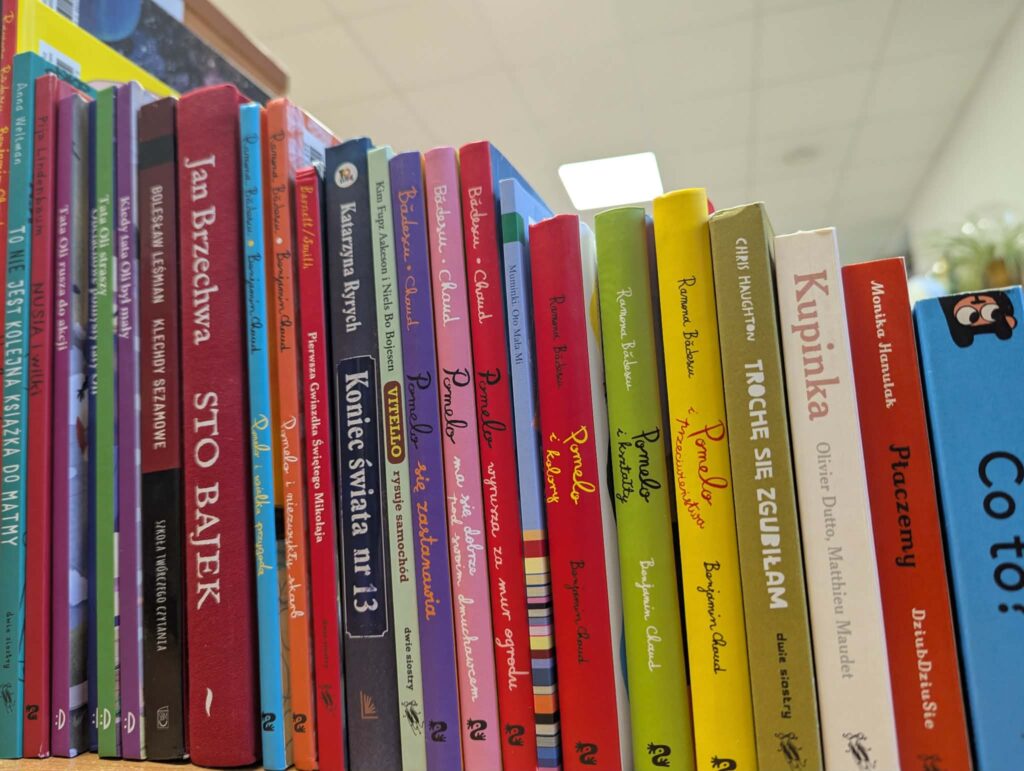 Na obrazie widoczne są kolorowe grzbiety książek ustawionych na półce. Książki mają różnorodne kolory i są ustawione pod kątem. Tło jest rozmyte, z widocznym biało-kremowym sufitem.