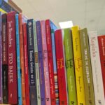 Na obrazie widoczne są kolorowe grzbiety książek ustawionych na półce. Książki mają różnorodne kolory i są ustawione pod kątem. Tło jest rozmyte, z widocznym biało-kremowym sufitem.