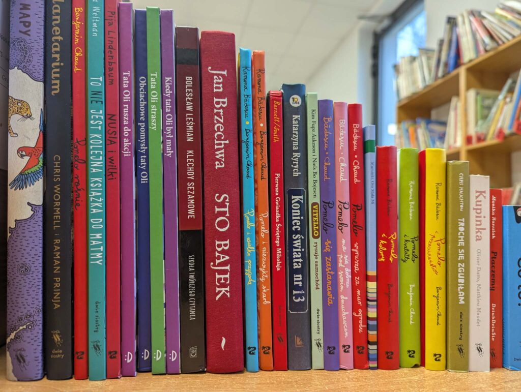 Kolorowe grzbiety książek stoją na półce w bibliotece. Tło to rozmyte regały i okno. Przyjemna atmosfera wiedzy i literatury.