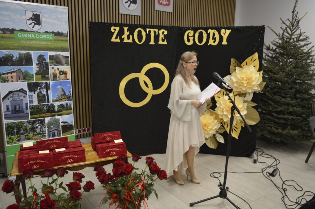 Kobieta w jasnej sukience przemawia przy mikrofonie na tle dekoracji „Złote Gody”. Po lewej stronie widać baner z krajobrazami gminy Dobre. Na stole stoją czerwone pudełka, otoczone bukietem czerwonych róż.