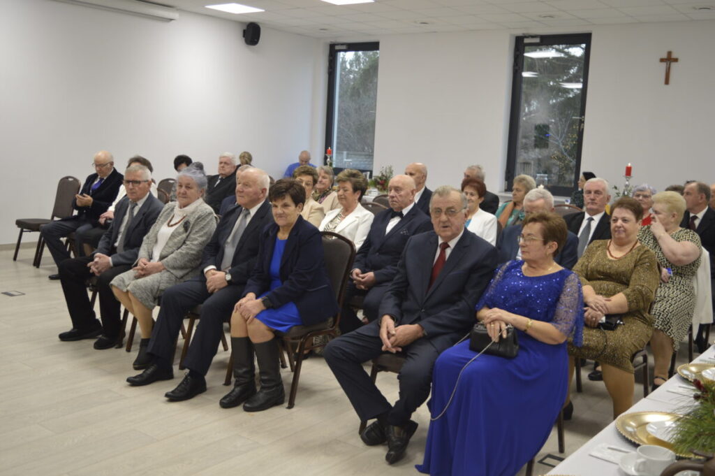 Na zdjęciu grupa elegancko ubranych osób siedzi w jasnej sali. Przeważają odcienie szarości, granatu i beżu. W tle widoczne są okna i krzyż na ścianie.