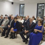 Na zdjęciu grupa elegancko ubranych osób siedzi w jasnej sali. Przeważają odcienie szarości, granatu i beżu. W tle widoczne są okna i krzyż na ścianie.