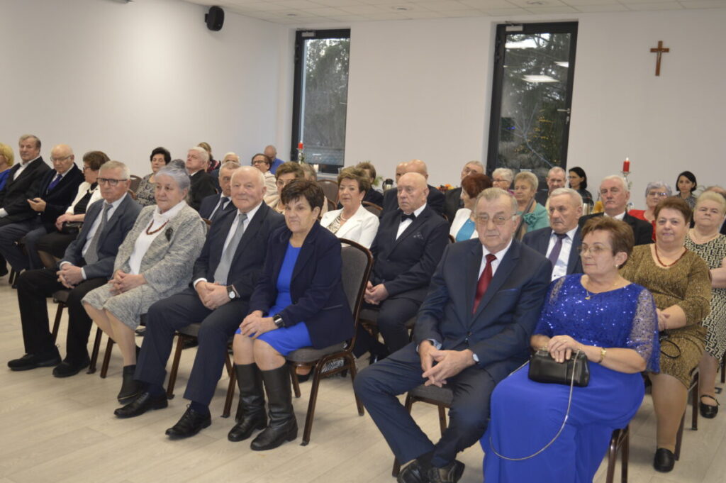 Grupa elegancko ubranych seniorów siedzi w jasnej sali. Na ścianie wisi krzyż. Przez okna widać drzewa i świece ozdobne.