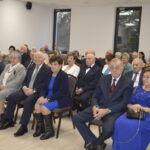 Grupa elegancko ubranych seniorów siedzi w jasnej sali. Na ścianie wisi krzyż. Przez okna widać drzewa i świece ozdobne.