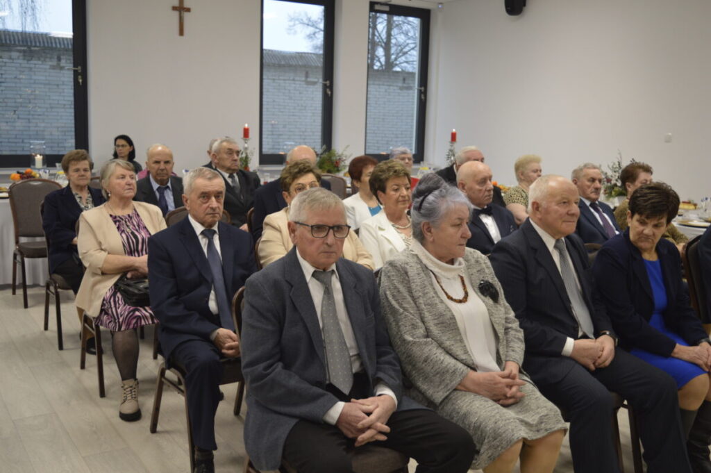 Grupa starszych osób w formalnych strojach siedzi na uroczystości w jasnym pomieszczeniu. W tle okna i ściana z krzyżem. Dekoracje stołów z owocami i świecami.