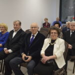 Grupa starszych osób siedzi w jasnym pokoju na krzesłach. Widać eleganckie stroje, dominują ciemne i jasne kolory. W tle są białe ściany i duże okno.