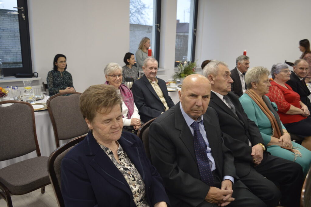 Grupa seniorów w eleganckich strojach siedzi w sali z białymi ścianami. Kolory dominujące to ciemne garnitury i czerwone ubrania. W tle widać okno i zastawiony stół.