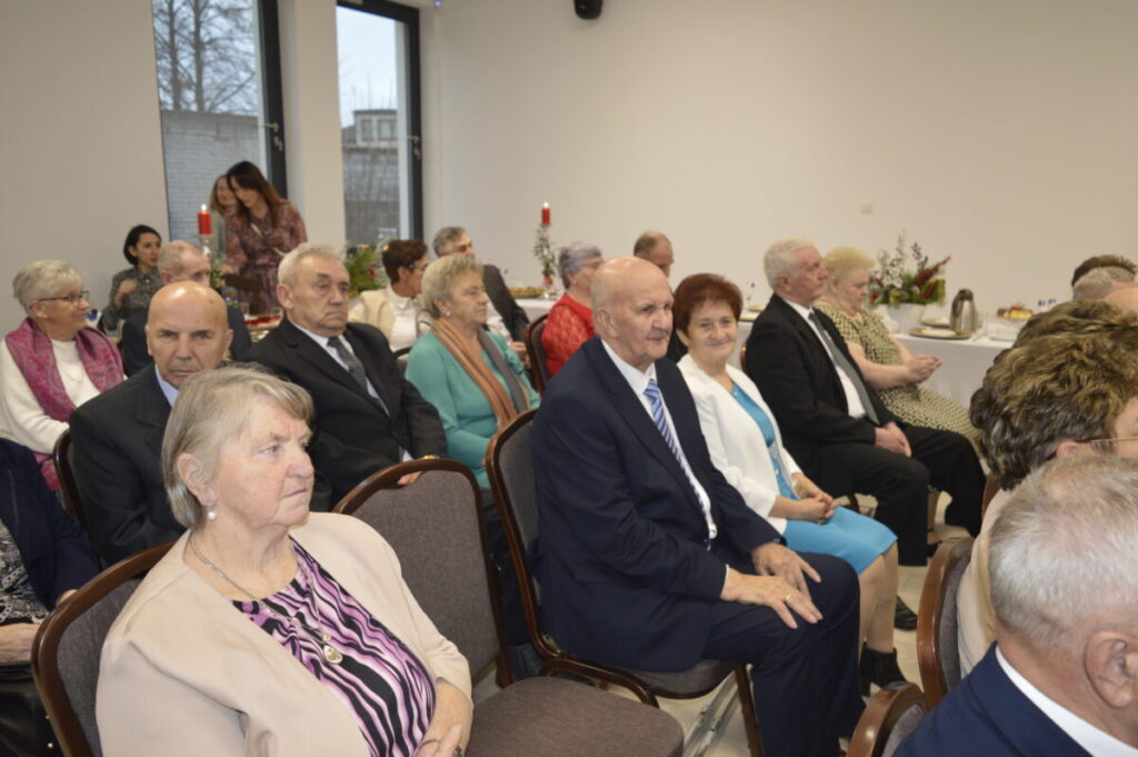 Grupa starszych osób siedzi na krzesłach w jasnym pomieszczeniu. Widoczne są eleganckie stroje: marynarki, białe bluzki. Na stole w tle stoją świeczniki i kwiaty.