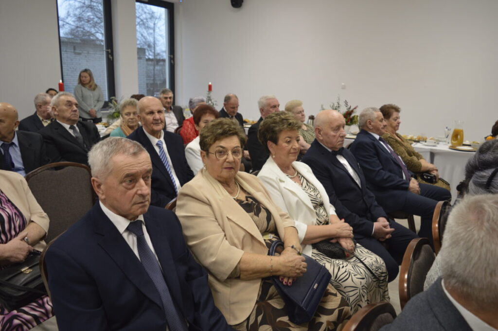 Grupa osób siedzi w jasnym pomieszczeniu podczas uroczystości. Dominują stroje formalne, a w tle widać okna i zastawione stoły. Dekoracje są świąteczne, z czerwonymi świecami na stołach.