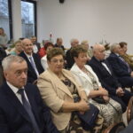 Grupa osób siedzi w jasnym pomieszczeniu podczas uroczystości. Dominują stroje formalne, a w tle widać okna i zastawione stoły. Dekoracje są świąteczne, z czerwonymi świecami na stołach.