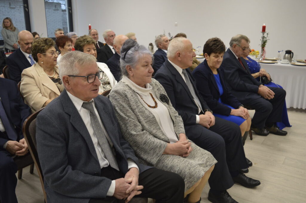 Na zdjęciu widać grupę starszych osób siedzących na krzesłach w jasnym pomieszczeniu. W tle ustawiony jest stół z białym obrusem. Kolory są stonowane, dominują szarości i granaty.