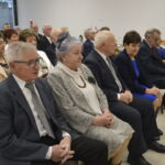 Na zdjęciu widać grupę starszych osób siedzących na krzesłach w jasnym pomieszczeniu. W tle ustawiony jest stół z białym obrusem. Kolory są stonowane, dominują szarości i granaty.