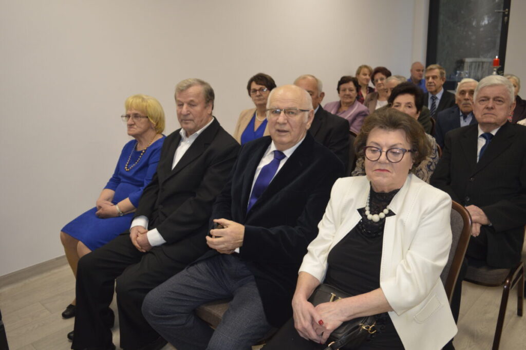 Grupa elegancko ubranych osób siedzi w pomieszczeniu z białymi ścianami. Kobieta na pierwszym planie nosi biały żakiet. W tle widoczne jest duże okno.