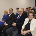 Grupa elegancko ubranych osób siedzi w pomieszczeniu z białymi ścianami. Kobieta na pierwszym planie nosi biały żakiet. W tle widoczne jest duże okno.