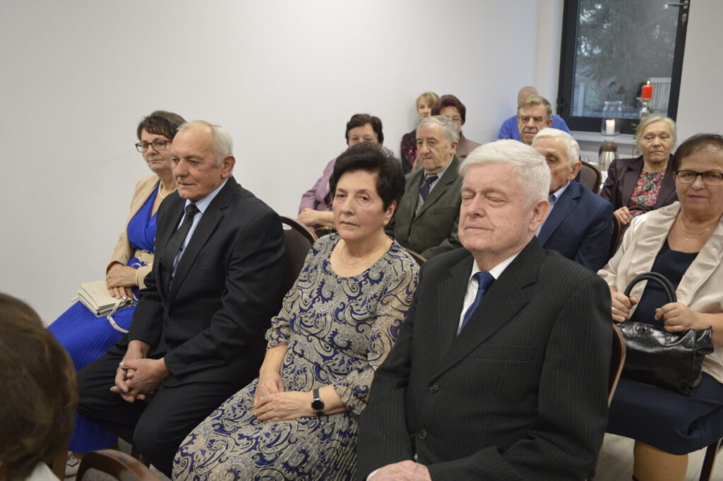 Grupa starszych osób siedzi na krzesłach w jasnym pomieszczeniu. Tło jest białe, a kilka osób ma na sobie eleganckie ubrania. W tle widać okno i zapaloną świecę.