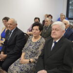 Grupa starszych osób siedzi na krzesłach w jasnym pomieszczeniu. Tło jest białe, a kilka osób ma na sobie eleganckie ubrania. W tle widać okno i zapaloną świecę.