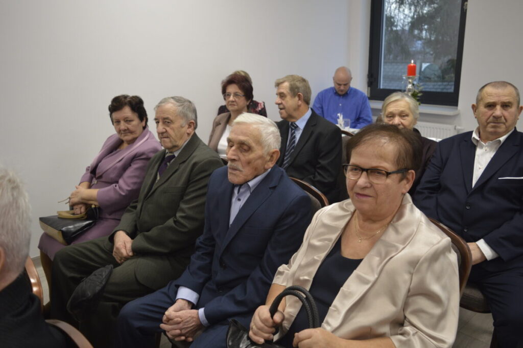 Grupa starszych osób siedzi w eleganckich strojach, na jasnym tle, z oknem i świecą w tle.