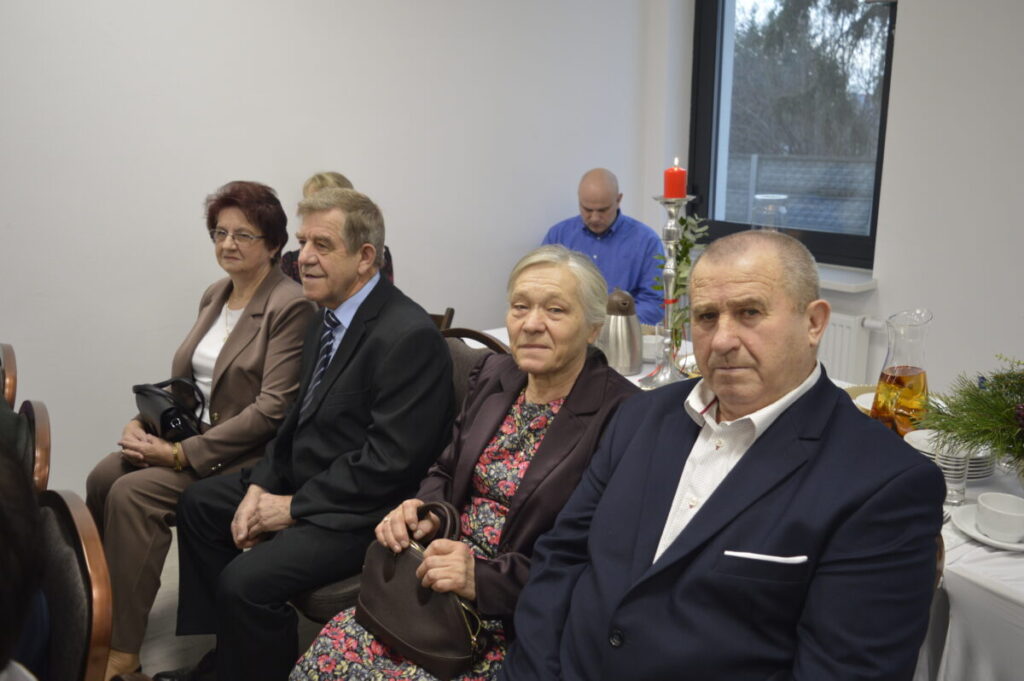 Na zdjęciu są cztery osoby siedzące w eleganckich strojach. Tło jest jasne z dużym oknem i dekoracją z czerwonej świecy. Obok znajdują się zastawa i dzbanek z herbatą.