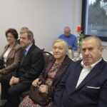 Na zdjęciu są cztery osoby siedzące w eleganckich strojach. Tło jest jasne z dużym oknem i dekoracją z czerwonej świecy. Obok znajdują się zastawa i dzbanek z herbatą.