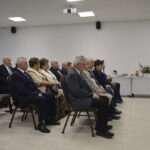 Grupa starszych osób siedzi na krzesłach w jasnej sali. Na ścianie biały projektor, a z boku stół z naczyniami. W tle jasne, prostokątne okna.