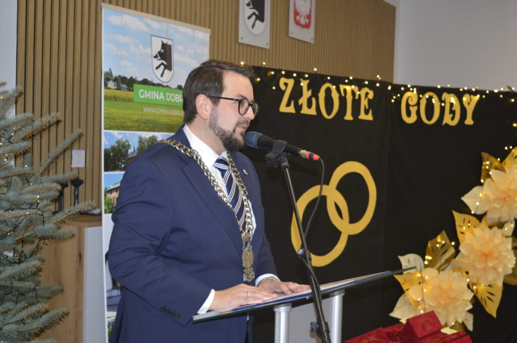 Mężczyzna w granatowym garniturze przemawia przy mikrofonie. W tle czarne tło z napisem ZŁOTE GODY i złotymi obręczami. Obok ozdoby kwiatowe i choinka.