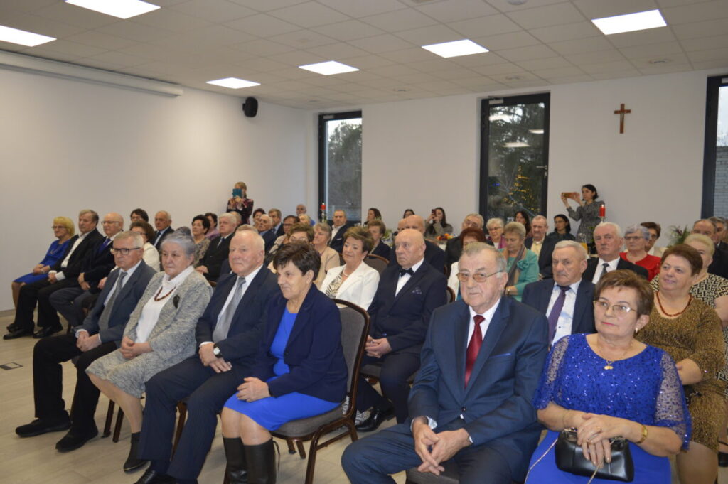 Grupa starszych osób w odświętnych strojach siedzi w jasnym pomieszczeniu z białymi ścianami i dużymi oknami. Na ścianie wisi krzyż. Tło jest neutralne, co pozwala skupić się na uczestnikach wydarzenia.