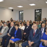 Grupa starszych osób w odświętnych strojach siedzi w jasnym pomieszczeniu z białymi ścianami i dużymi oknami. Na ścianie wisi krzyż. Tło jest neutralne, co pozwala skupić się na uczestnikach wydarzenia.