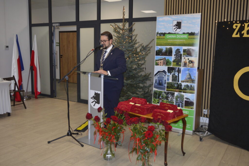 Na zdjęciu mężczyzna przy mównicy, za nim choinka i plakaty. Obrus jest bordowy, wazon z czerwonymi różami stoi obok. Tło jest jasne, widoczne flagi.