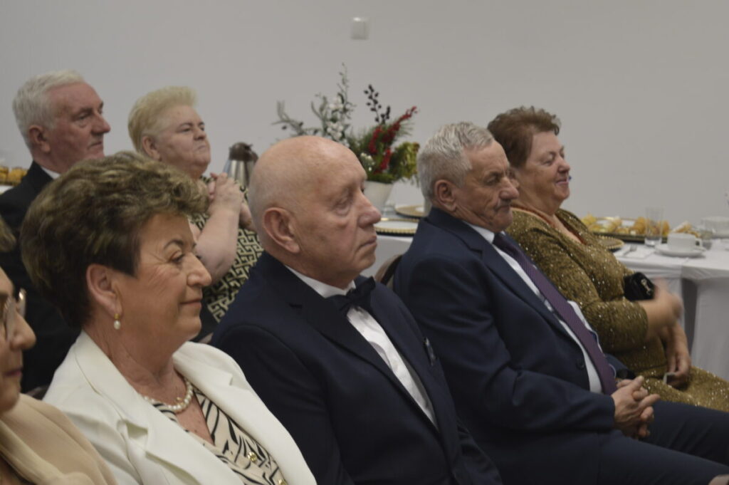 Grupa starszych osób siedzi przy stole w eleganckim stroju. W tle biały pokój z dekoracjami i zastawą stołową. Atmosfera formalna i uroczysta.