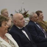 Grupa starszych osób siedzi przy stole w eleganckim stroju. W tle biały pokój z dekoracjami i zastawą stołową. Atmosfera formalna i uroczysta.