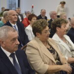 Grupa elegancko ubranych osób siedzi w sali. W tle widać białe ściany i świąteczne dekoracje. Atmosfera formalna i skupiona.