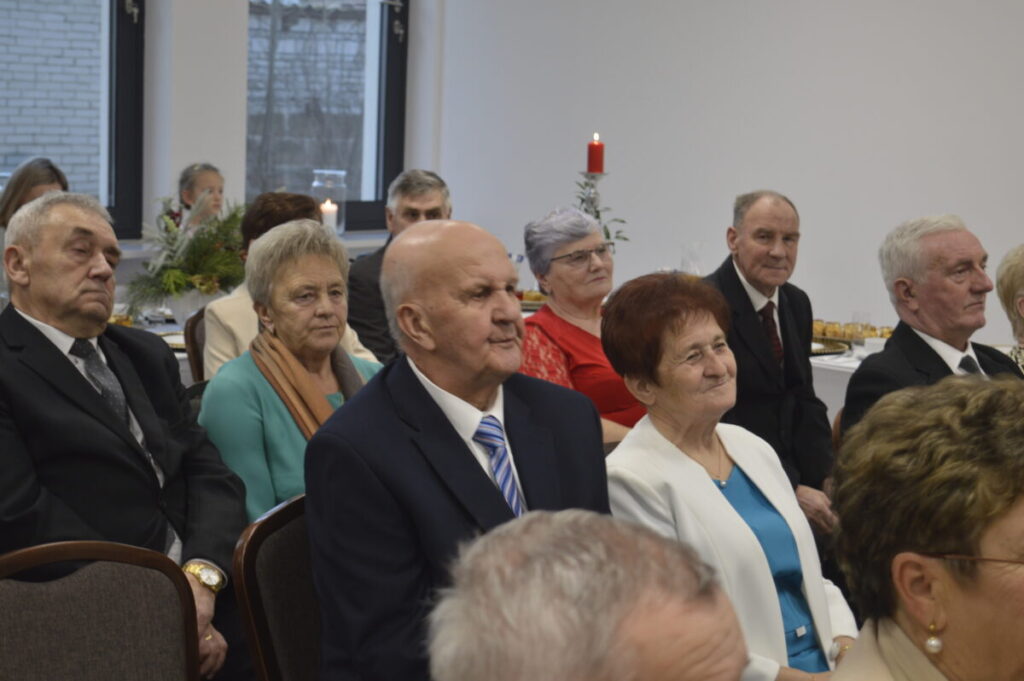 Grupa starszych osób siedzi w pomieszczeniu. Tło jest jasne z dekoracjami i świecami. Wszyscy są elegancko ubrani.
