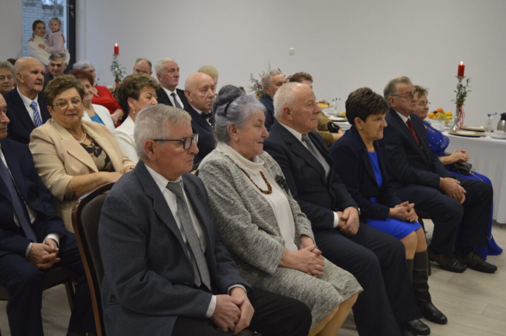 Grupa elegancko ubranych starszych osób siedzi na uroczystości. W tle biały stół z dekoracjami i czerwonymi świecami. Atmosfera oficjalna.