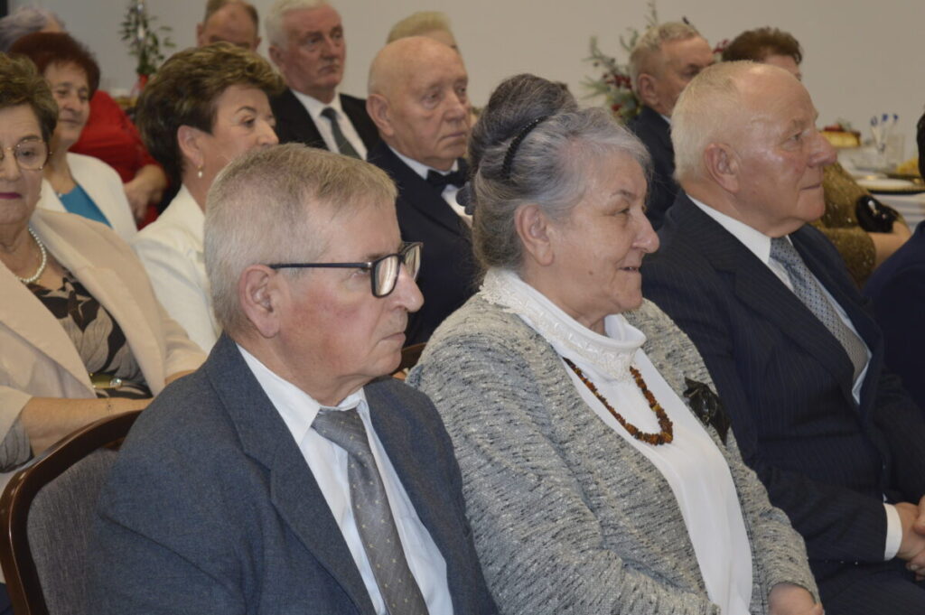 Grupa starszych osób siedzi w rzędzie w eleganckich strojach. Dominują odcienie szarości i niebieskiego. W tle widać elementy dekoracyjne.