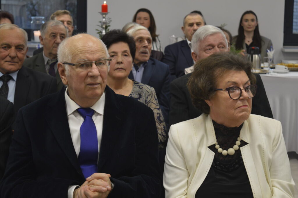 Oto grupa starszych osób siedzących razem na uroczystości. Widoczne eleganckie ubrania, biały obrus w tle. Czerwona świeca dodaje świątecznego nastroju.