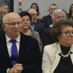 Oto grupa starszych osób siedzących razem na uroczystości. Widoczne eleganckie ubrania, biały obrus w tle. Czerwona świeca dodaje świątecznego nastroju.