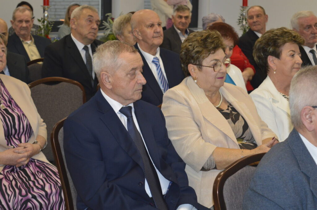 Grupa starszych osób siedzi na sali. Tło jest jasne, z czerwonymi świecami. Dominują kolory niebieski, beżowy i czarny.