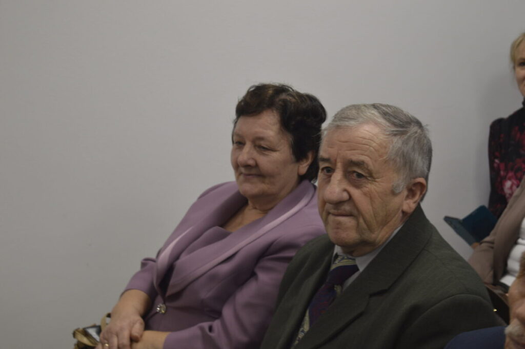 Nie znam tych osób, ale zdjęcie przedstawia dwie starsze osoby siedzące w pomieszczeniu o białych ścianach. Kobieta w liliowym kostiumie, mężczyzna w ciemnej marynarce. Obok widoczna inna osoba.