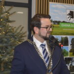 Mężczyzna w garniturze przemawia przy mikrofonie obok choinki z lampkami. W tle plakat z nazwą Gmina Dobre.