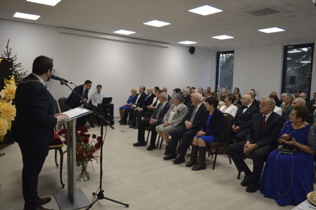 W sali z jasnymi ścianami i drewnianą podłogą mężczyzna przy mównicy przemawia do siedzącej publiczności. Po lewej duże żółte kwiaty, w tle choinka. Uczestnicy są elegancko ubrani.