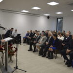 W sali z jasnymi ścianami i drewnianą podłogą mężczyzna przy mównicy przemawia do siedzącej publiczności. Po lewej duże żółte kwiaty, w tle choinka. Uczestnicy są elegancko ubrani.