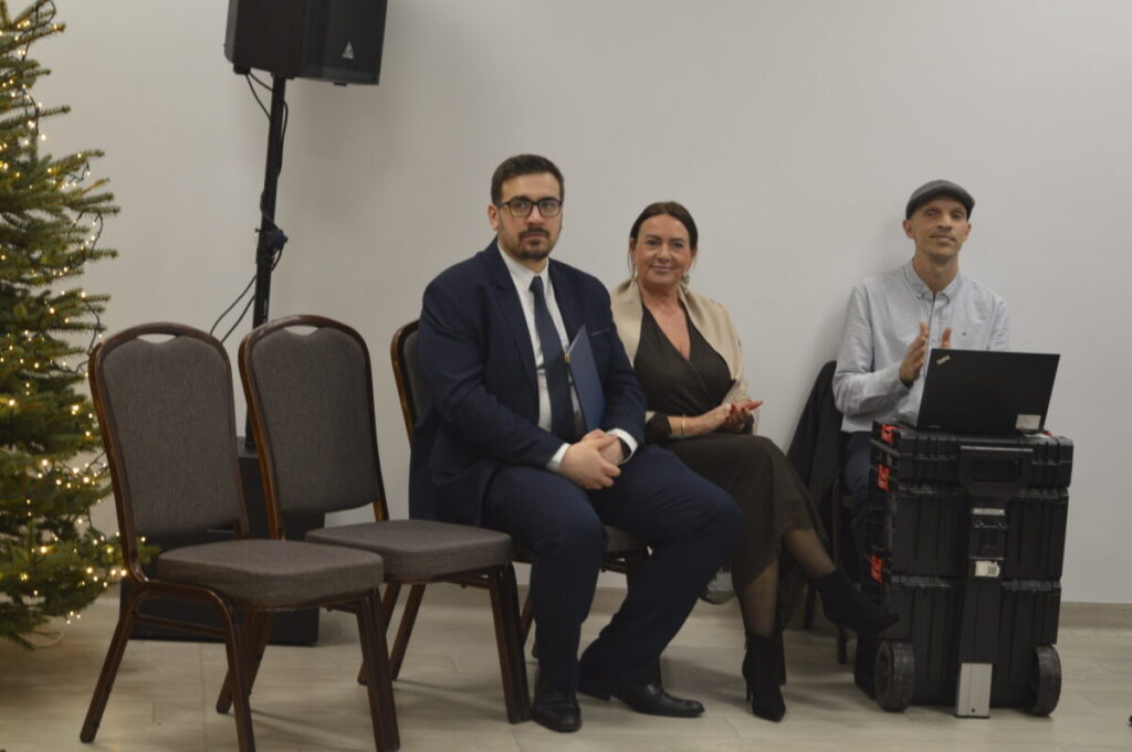 Trzy osoby siedzą na krzesłach; mężczyzna w garniturze, kobieta w sukni, trzeci mężczyzna z laptopem; obok choinka i białe tło.