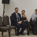 Trzy osoby siedzą na krzesłach; mężczyzna w garniturze, kobieta w sukni, trzeci mężczyzna z laptopem; obok choinka i białe tło.