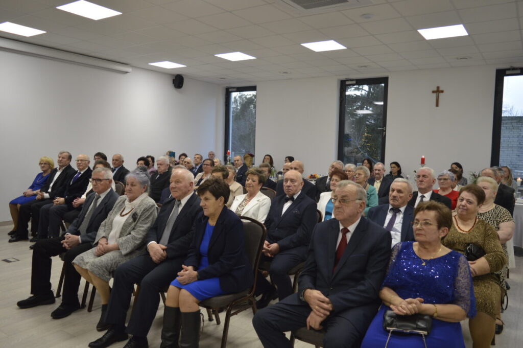 Ludzie w eleganckich ubraniach siedzą w jasnej sali, na ścianie krzyż, za oknami drzewa. Dominuje kolor granatowy i biały.