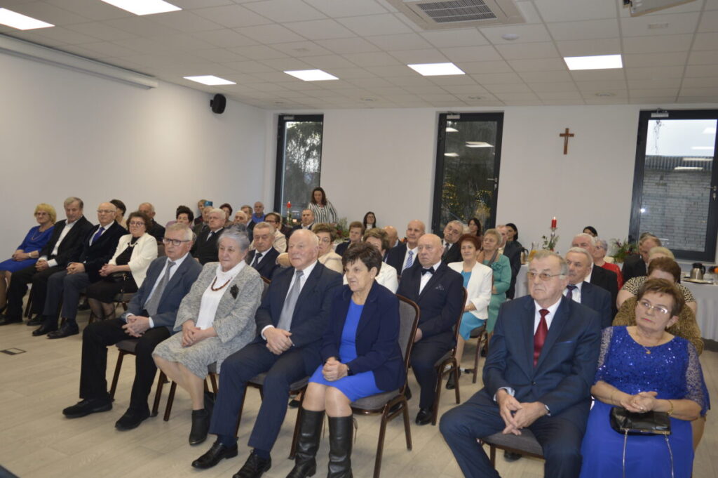 Grupa starszych osób siedzi w jasnej sali konferencyjnej. W tle białe ściany z krzyżem, okna z ciemnymi ramami. Osoby są elegancko ubrane.